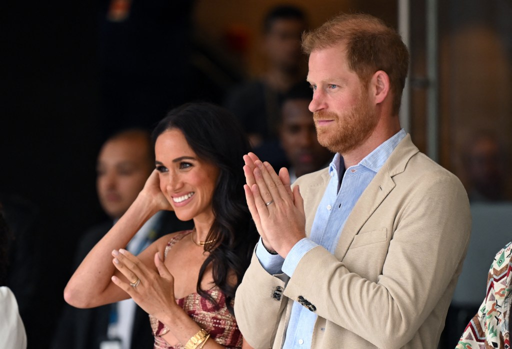 Meghan Markle, in a patterned summer dress, stands next to Prince Harry, in a cream blazer and blue shirt.